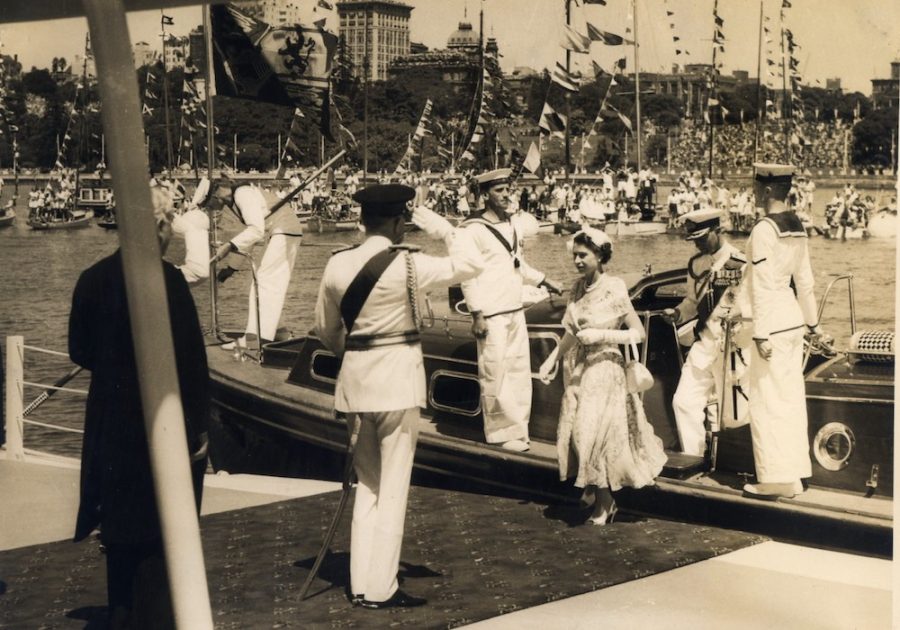 Queen Elizabeth steps off a boat onto Sydney Harbour. Prince Philip follows her. A man in military attire salutes her. Men in naval attire flank her.