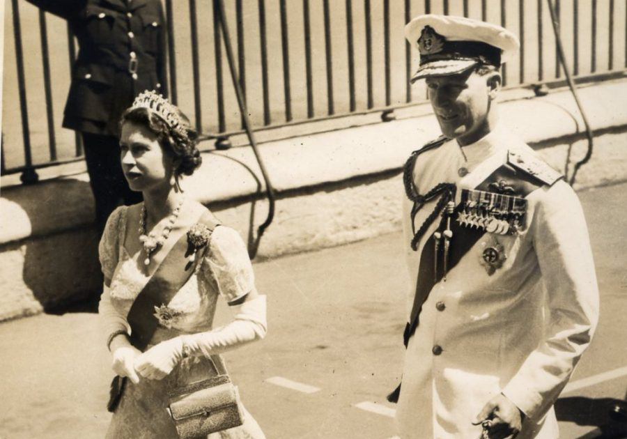 Queen Elizabeth walks with Prince Philip. She is wearing in formal regalia and a tiara crown. He is in naval attire.