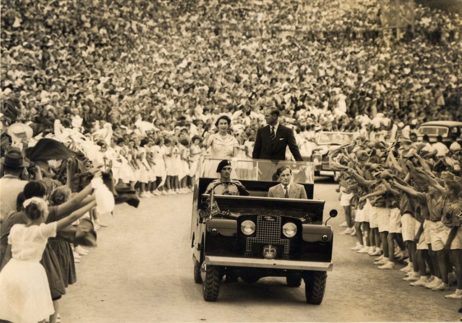 Queen Elizabeth stands in roofless car and waves to crowds of people as she passes by.