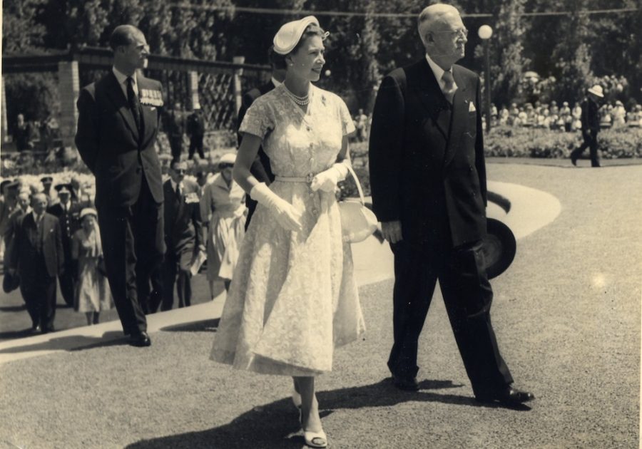 Queen Elizabeth is shown around the Botanic Gardens. She wears a white lace dress and white gloves. Prince Philip follows behind her.