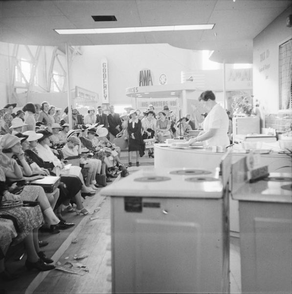 A women conducts a cooking show. She is watched by a crowd of seated women.