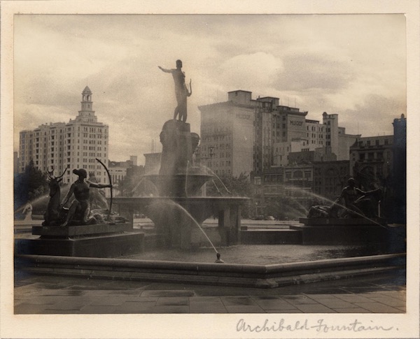 Black and white photograph of the Archibald Fountain in Sydney. Statues of Greek gods and goddesses are seated around the fountain. In the background are the city buildings.