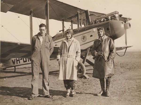 A man in overalls stands beside a woman dressed in a woollen overcoat and wearing a leather aviation cap and goggles. She smiles at the camera while putting her hands in her pockets. Beside her is another man dressed in a leather overcoat. He is also wearing an aviation cap and goggles and is holding a pair of leather gloves. In the background is a single-engine aeroplane.