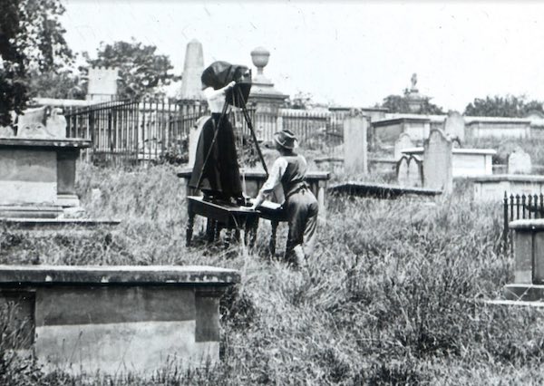 The pair are standing in the middle of the Devonshire Street Cemetery and are surrounded by graves and headstones. Mrs Foster stands on a table and adjusts a camera from underneath a hood. A man in a brimmed hat holds the table steady.