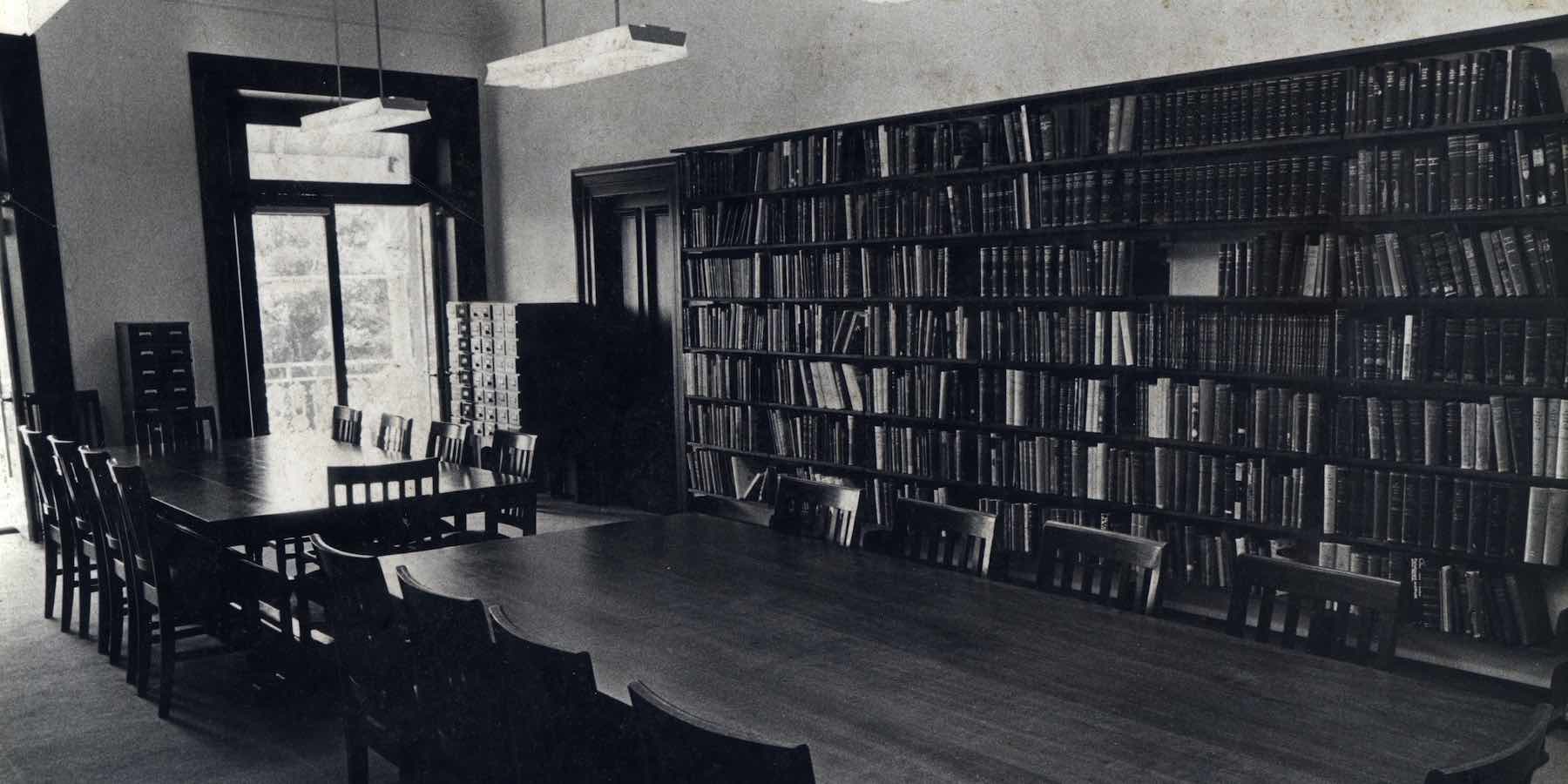 In the library's centre are two long wooden tables with wooden chairs. The back wall features seven shelves that are filled with books. A window and a set of archive drawers are in the corner of the room.