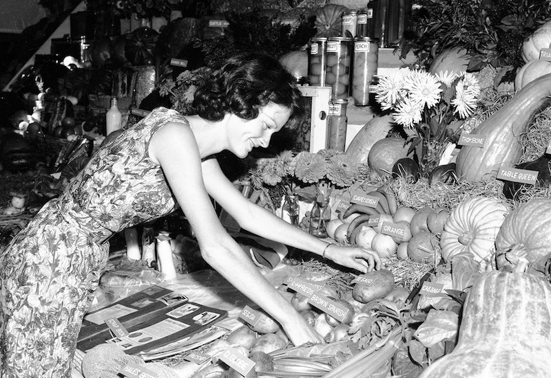 Black and white photograph of a woman smiling while she inspects the labels produce display. Various vegetables are on display with identifying labels.