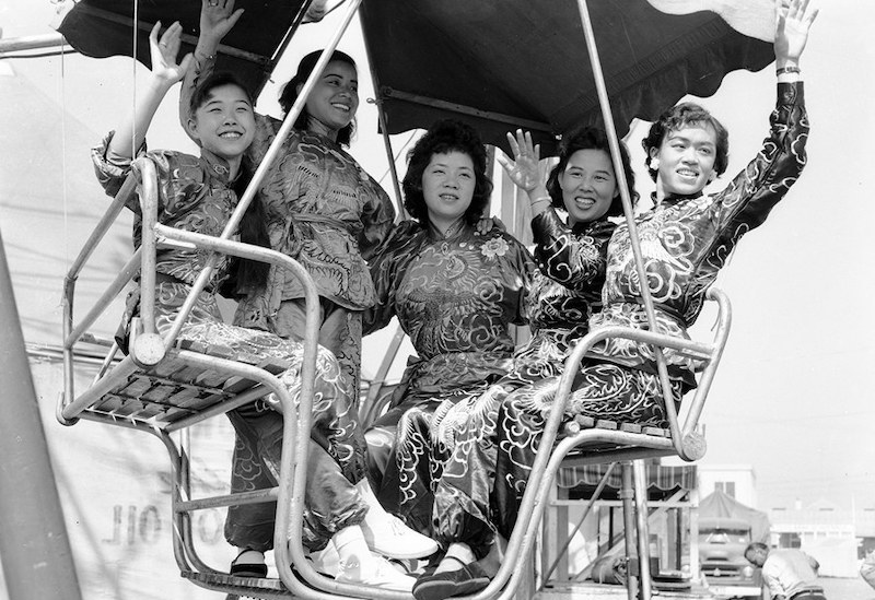 Black and white photograph of five Asian people, four women and a younger man, all wearing traditional South-East Asian attire. They are waving and smiling while they ride on the Ferris wheel.