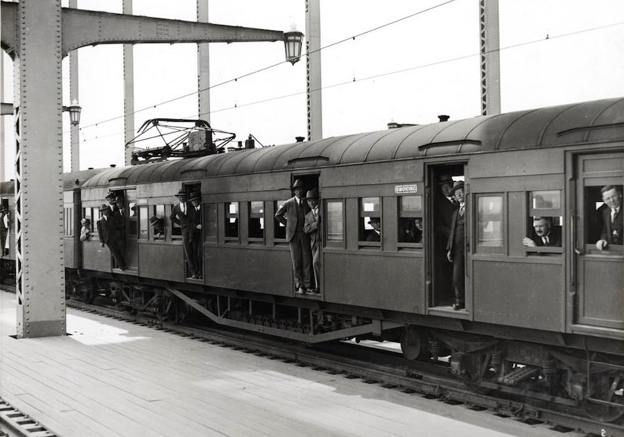Men wearing three-piece suits and bowler hats stand at the edge of the train doors and look out the windows as it pulls into a station.