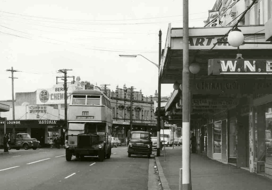 A black and white photograph of a double-decker bus travelling along the street.