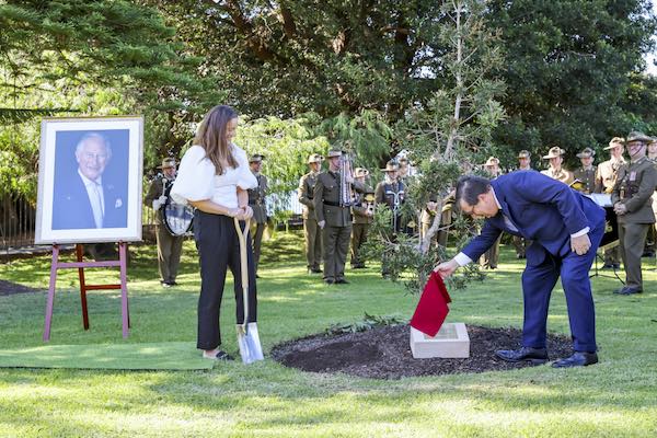A woman stands beside a portrait of King Charles while holding a shovel. A man removes a red covering from a stone plaque in front of a tree sapling. Men in military uniform stand in the background.