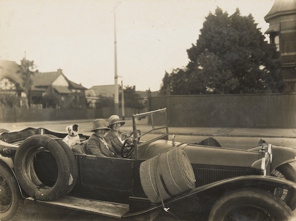 Jean Robertson and Kathleen Howell seated in Lancia Lambda with Barney the dog, car parked in suburban street.