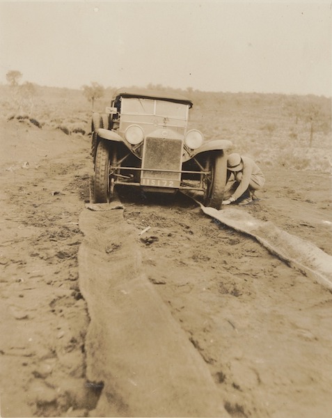 Looking along matting placed under front wheels of the Lancia, Kathleen Howell bent down placing matting under front wheel, wide flat country in background.