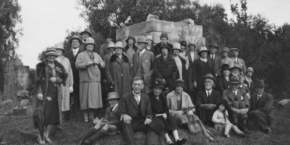 A large group of well-dressed men and women pose for a photograph at a sandstone memorial.