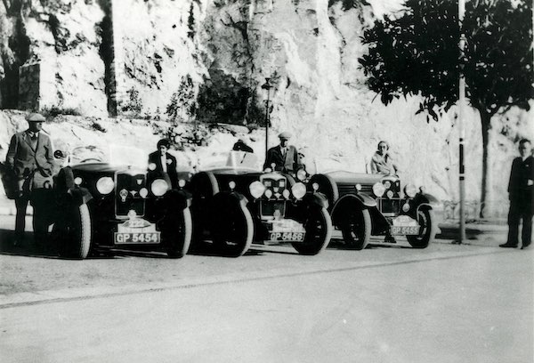 Three vehicles parked in front of cliff face, a man or woman standing beside each car, woman on far right, Joan Richmond.