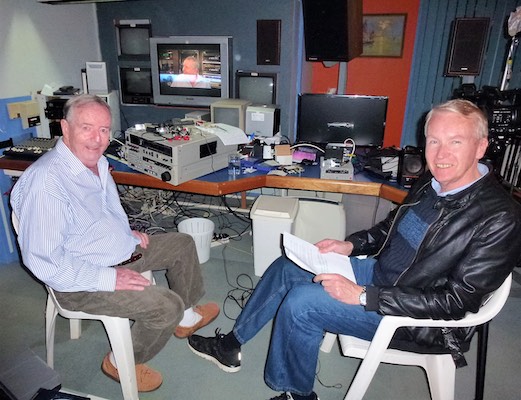 Graham Shirley and Dick Collingridge look and smile at the camera. Graham holds a paper script in his hand. A variety of audio-visual technology is in the background.