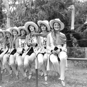 A black and white photograph of seven women dressed as cowgirls sitting on a fence. An ostrich stands behind the fence.