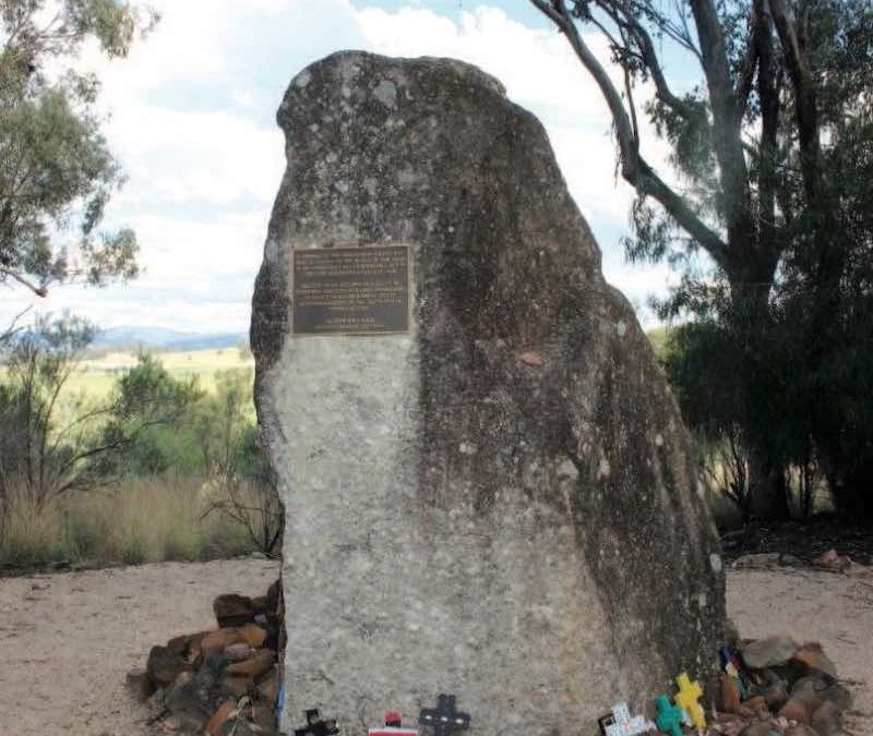 A photograph of Memorial Rock at the Myall Creek Massacre Site. A plaque is fixed onto the rock. Laid at the rock's base are small crucifixes.