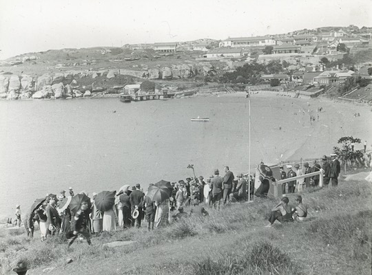 A black and white photograph showing a group of people standing on a hill above the beach at Camp Cove. They are holding parasols and are well dressed.
