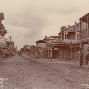 A sepia photograph looking south of Mann Street in Gosford. A dirt road is in the centre and buildings line the street. Signs read 'Hairdresser', 'Tobacconist', and 'Royal Hotel'. Horse-drawn carts are visible.