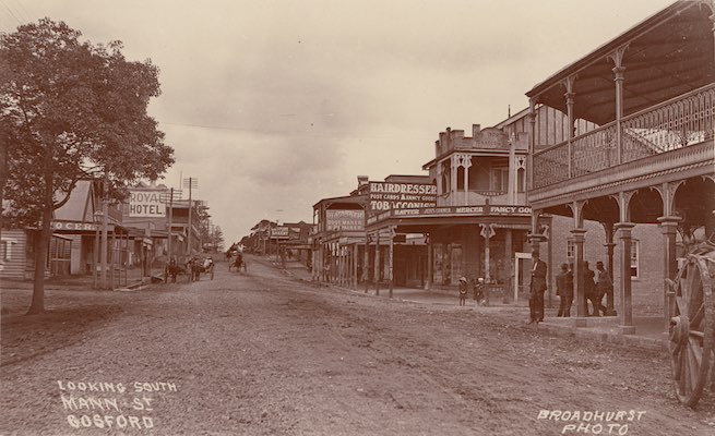 A sepia photograph looking south of Mann Street in Gosford. A dirt road is in the centre and buildings line the street. Signs read 'Hairdresser', 'Tobacconist', and 'Royal Hotel'. Horse-drawn carts are visible.