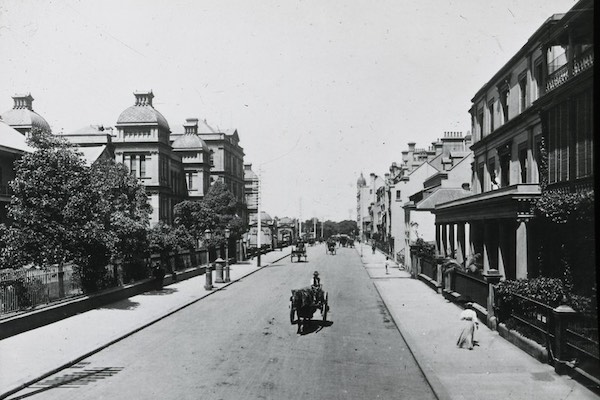 Black and white photograph of Macquarie Street, Sydney, circa early 1900s. A horse-drawn cart travels down the centre of a wide, quiet street lined with grand buildings, iron fences, and leafy trees. Ornate domed rooftops are visible on the left, while verandah-fronted terraces stand to the right. A few pedestrians are seen walking on the footpaths.