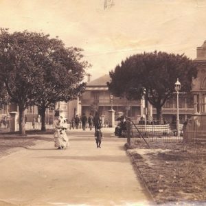 A sepia photograph showing the Frazer Fountain in Hyde Park, Sydney. A woman in a crinoline dress and sun hat, holding a baby, walks by, accompanied by a boy who is looking at the fountain.