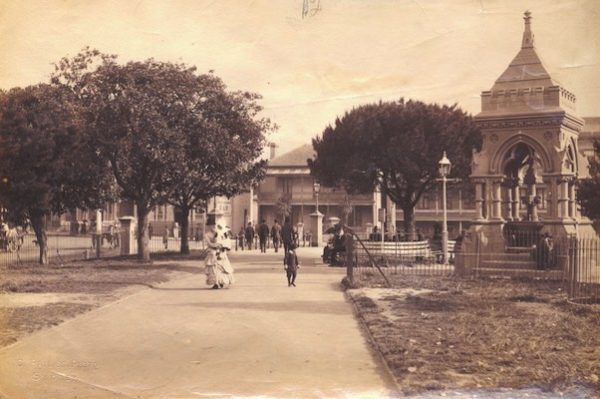 A sepia photograph showing the Frazer Fountain in Hyde Park, Sydney. A woman in a crinoline dress and sun hat, holding a baby, walks by, accompanied by a boy who is looking at the fountain.