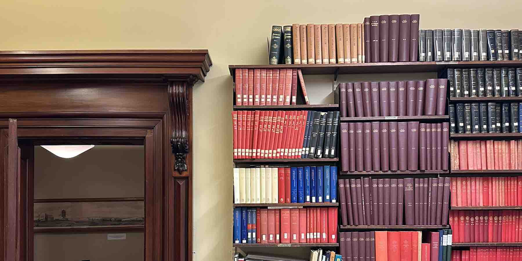 Bookshelves in the RAHS Library filled with bound historical journals in red, maroon, blue, and black covers beside a carved timber doorway