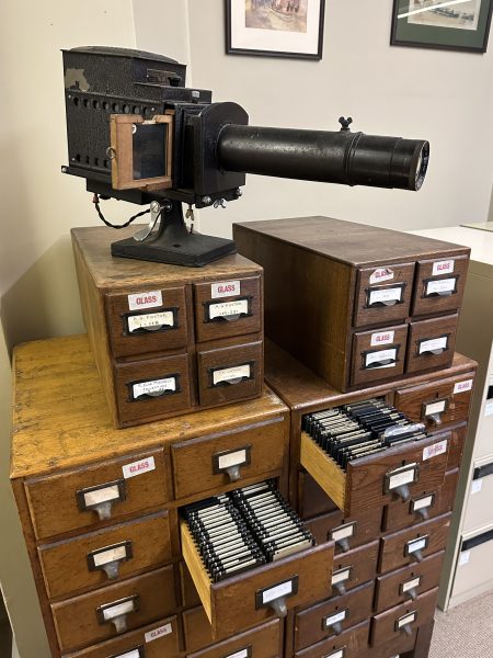 Photograph of the RAHS glass slide collection and original lantern projector. Several rectangular glass slides are displayed on a white cloth alongside the vintage metal projector, which was used by RAHS members in the early 20th century to deliver illustrated lectures on Australian history