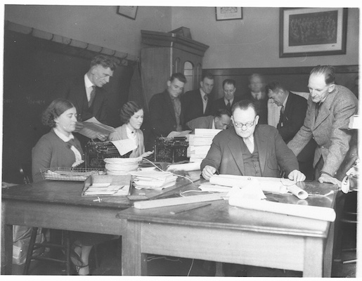 A photograph showing a group of men huddled over documents on a desk, while two women use typewriters.