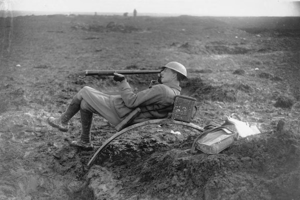 A photograph of Charles Bean lying in muddy trenches and using a telescope. Bean's boots are caked with mud, and he is wearing a Brodie helmet.
