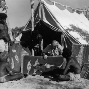 A photograph of four Aboriginal men and a person of European descent looking at bark paintings. A makeshift tent is in the background.