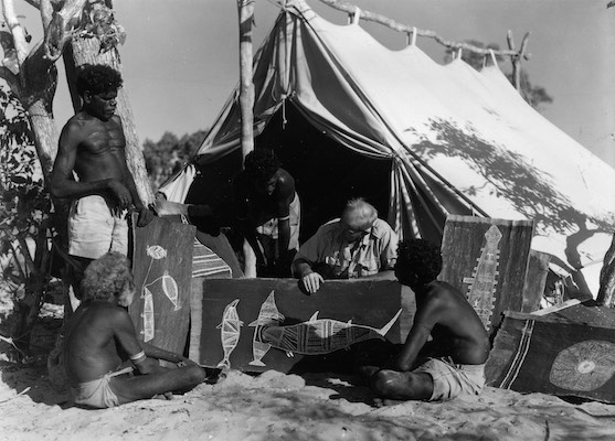 A photograph of four Aboriginal men and a person of European descent looking at bark paintings. A makeshift tent is in the background.