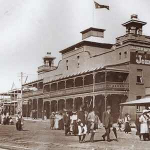 A sepia photograph showing the Brighton Hotel in Kiama in 1910. An Australian flag flies above the hotel. Groups of families walk along a dirt road in the foreground.