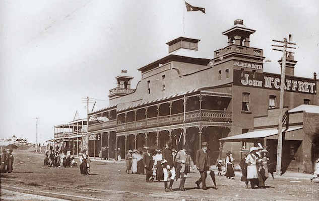 A sepia photograph showing the Brighton Hotel in Kiama in 1910. An Australian flag flies above the hotel. Groups of families walk along a dirt road in the foreground.