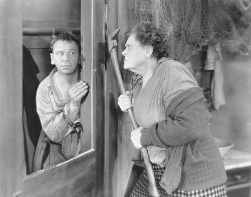 A screenshot taken of the film Min and Bill. The image shows an angry woman holding an axe and staring at a man.