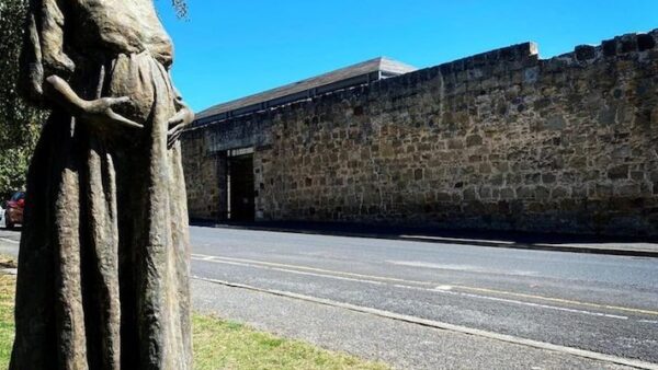 In the foreground is a statue of a visible pregnant female convict. The stone wall of the Cascades Female Factory is in the background.
